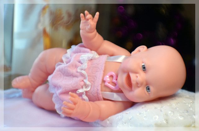 toy smiling no teeth and wearing a little pink dress and heart necklace, laying in bed sideways while hands are up and twisting a bit, white pillow underneath