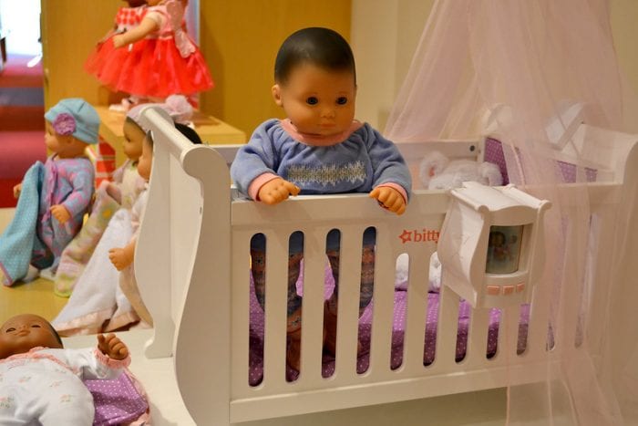 American Girl Bitty is popular in the USA. Very realistic looking baby doll in a crib wearing a blue and pink sweater staring and standing up from the crib. Other little kid toys surrounding 