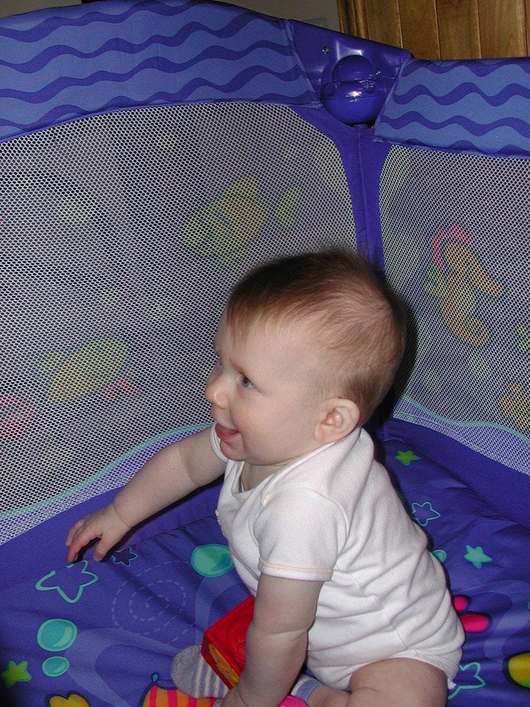 A smiling toddler inside his blue Nuna crib, relishing in a safe environment as he explores his surroundings filled with toys.