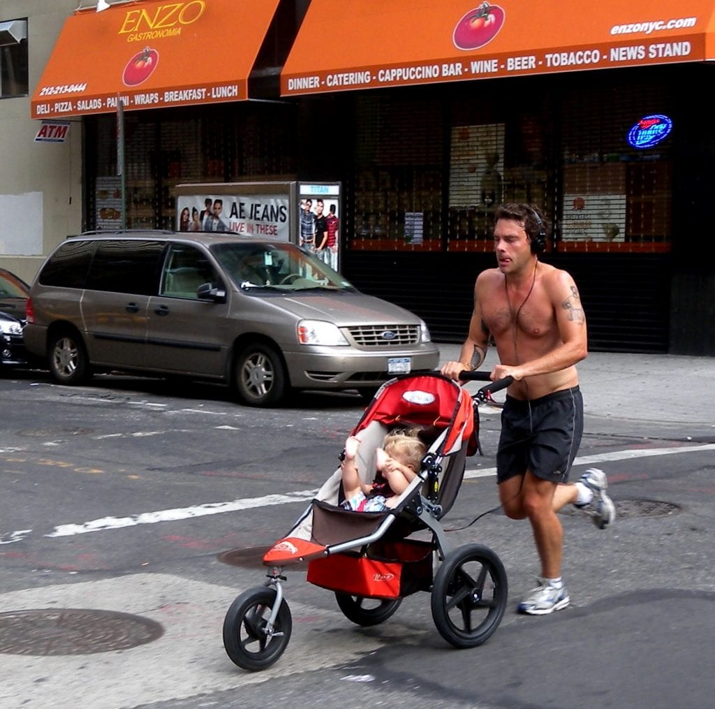 Select Stroller: A jogger pushes a baby in a red jogging stroller through a bustling city street, with storefronts and a minivan in the background.