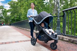 A content baby sits in a B-Ready stroller, pushed by a smiling person on a tree-lined, brick-paved path.