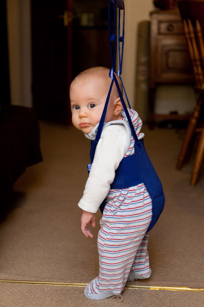 A baby wearing a striped one piece rides one jump toy available on the market while in the dim living room.