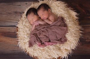Twin infants are nestled together in serene sleep, swaddled gently in a plush brown blanket placed within a cozy, cream-colored, furry basket. They rest against the rich contrast of a dark wooden floor, symbolizing warmth and tranquility. This tender scene embodies the pure, silent bond shared between siblings, creating a picture of peaceful repose and familial intimacy.