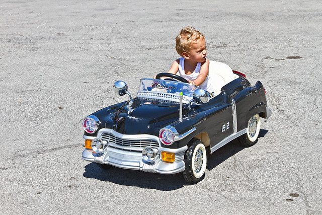 A kid riding a classic vintage black pedal car in the outside pavement