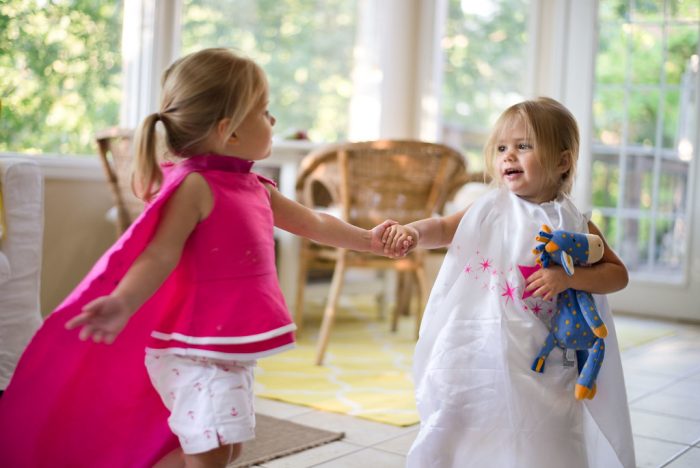 A pair of adorable young girls, one draped in a delicate pink attire, while the other sports an elegant white outfit and is holding a stuffed toy. The contrast in their dresses adds to the scene's color palette - presenting a visual treat with hues symbolizing innocence and charm typical of this tender age group. Their outfits may differ, but their camaraderie and playful spirit unify them on this exciting day full of joyous laughter and endless giggles.