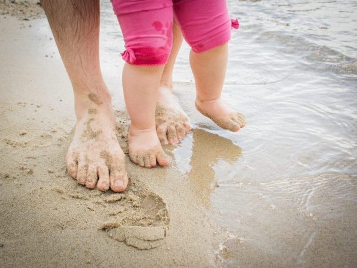 support for shoes - A father supporting baby's ankle as they practice walking on the sand without shoes.
