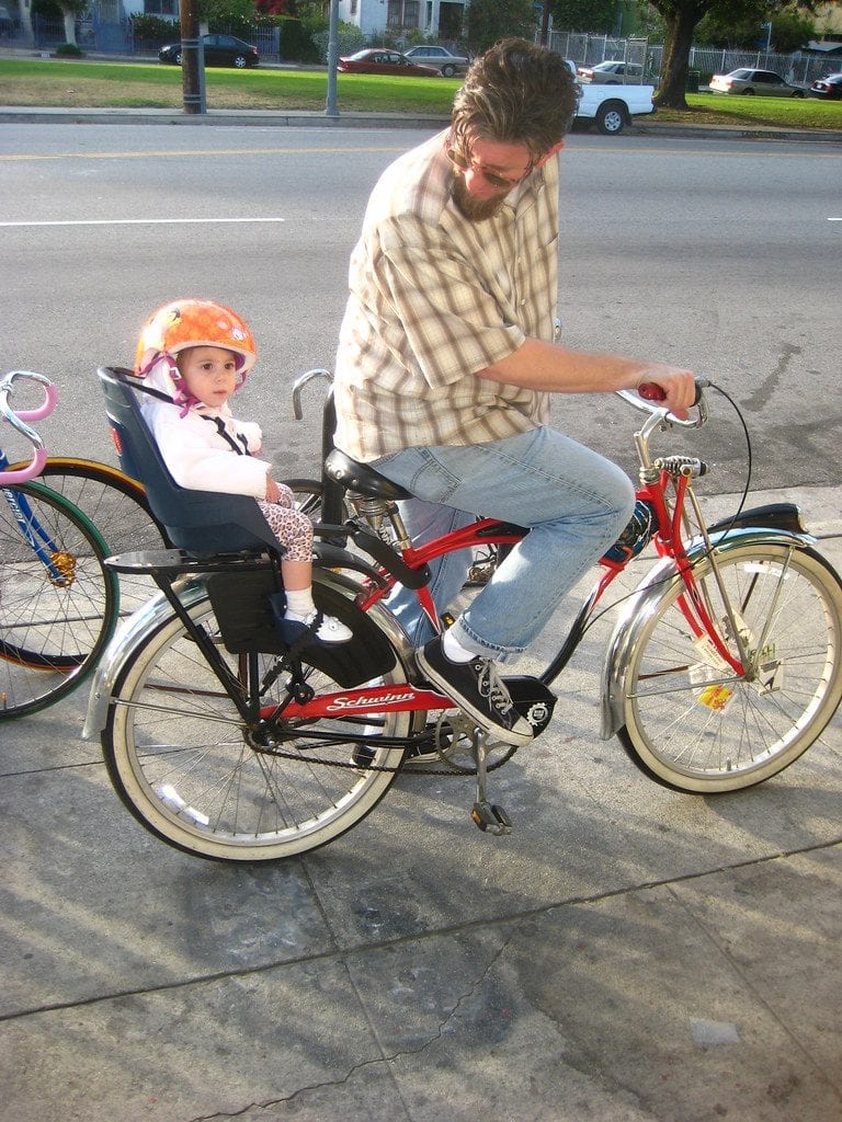 A father riding a bicycle watching her daughter setting at the baby carrier attached on the bike. 