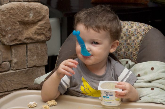 The baby sits on a comfortable baby chair in the kitchen while eating his yogurt.