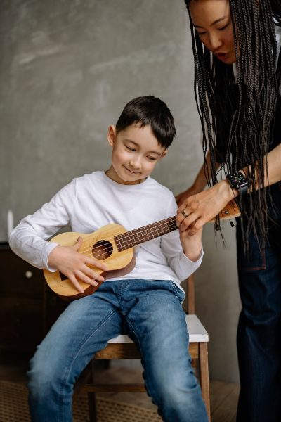 In a warmly lit room, a devoted mother is seated beside her son, their shared focus on a vibrant musical journey. The young boy, eager to learn, cradles a guitar in his hands, while his mother, with a patient and encouraging demeanor, guides him through the intricacies of playing. The soft glow of natural light bathes them in a comforting atmosphere, underscoring the tenderness and bond between parent and child. Together, they embark on a journey of discovery and skill development, as the melodious notes of the guitar weave through the air, echoing the harmonious connection between a mother and her son in the shared pursuit of artistic expression and joyous learning.