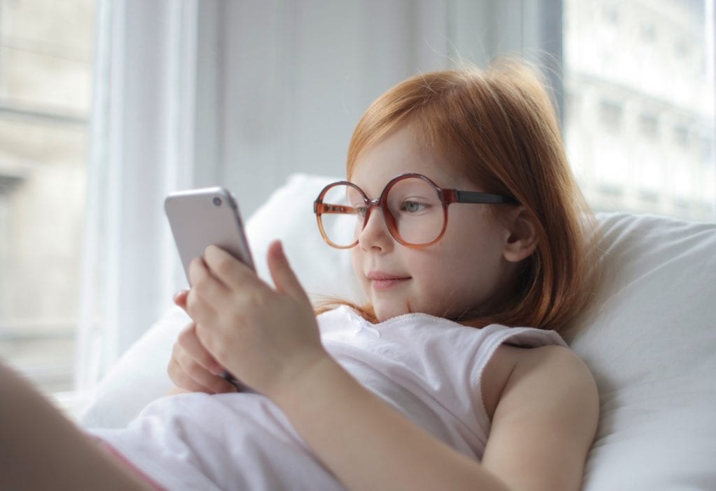 A little girl with eyeglasses happily playing with her cellphone on bed. It's a challenge!