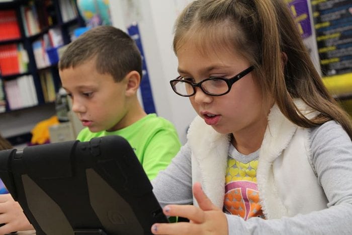 Two kids sit together in the room while playing with their iPad. 