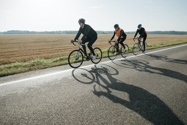 Three bicyclists riding in a line on a secondary road with a field in the background
