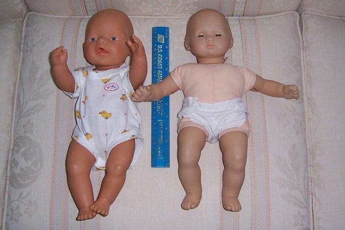 There are two infant dolls. The one looks like he's asleep and the other is not. They are wearing different clothes. 