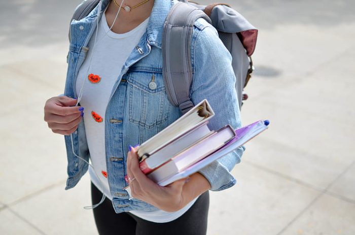 A girl wearing denim jacket is holding books and a backpack.