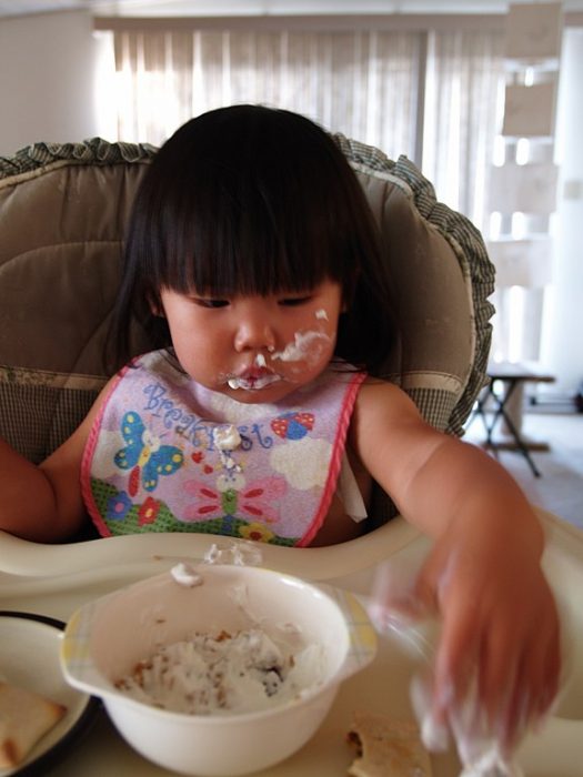 The baby sits on her best chair while eating a yogurt for babies.