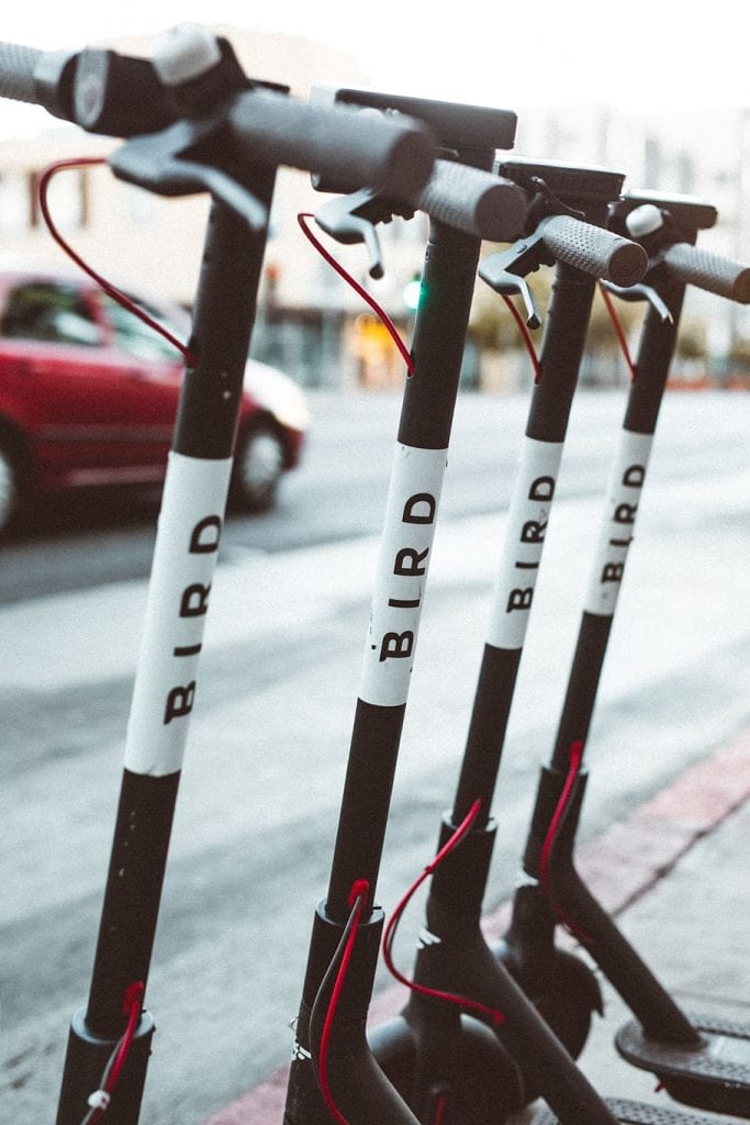 A row of four Bird electric scooters parked on a sidewalk. They are all black with white Bird logos on the front and sides. The handlebars are turned to the left, and the wheels are locked. 