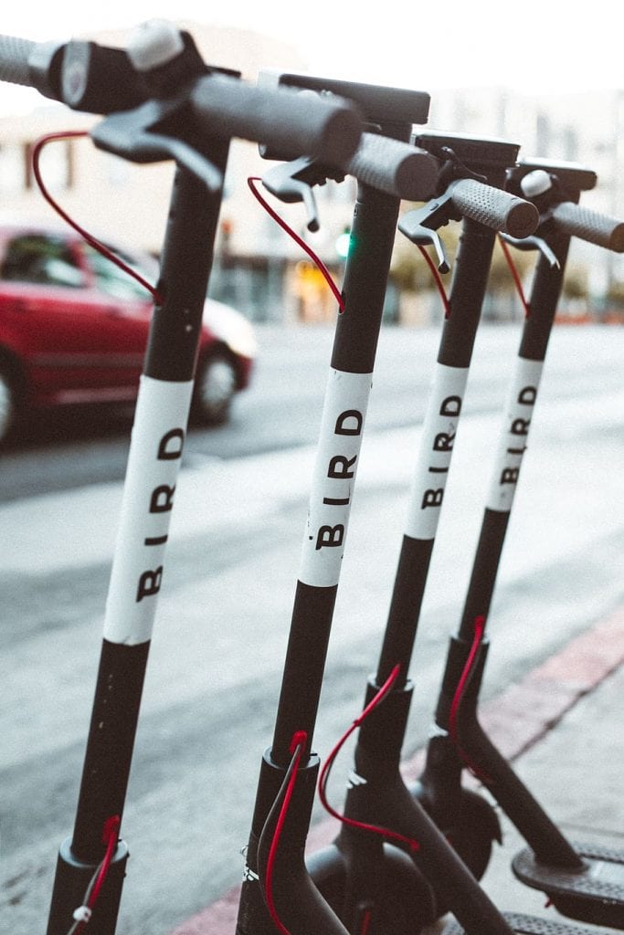 A row of four Bird electric scooters parked on a sidewalk. They are all black with white Bird logos on the front and sides. The handlebars are turned to the left, and the wheels are locked.