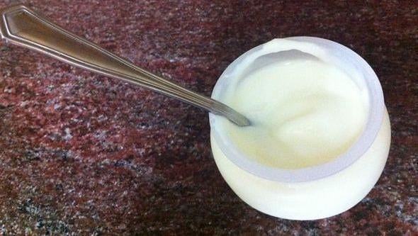 A glass jar of yogurt with a metal spoon sticking out of it. The yogurt is plain white and sits in a clear glass jar. The spoon is resting upright in the center of the yogurt, with a small amount of yogurt clinging to the sides. The jar is sitting on a light-colored wooden table, and there is a white napkin or paper towel to the left of the jar.The photo is taken from a slightly elevated angle, so we can see the top of the yogurt and the entire spoon. The background is blurred, so the focus is on the yogurt and spoon.
