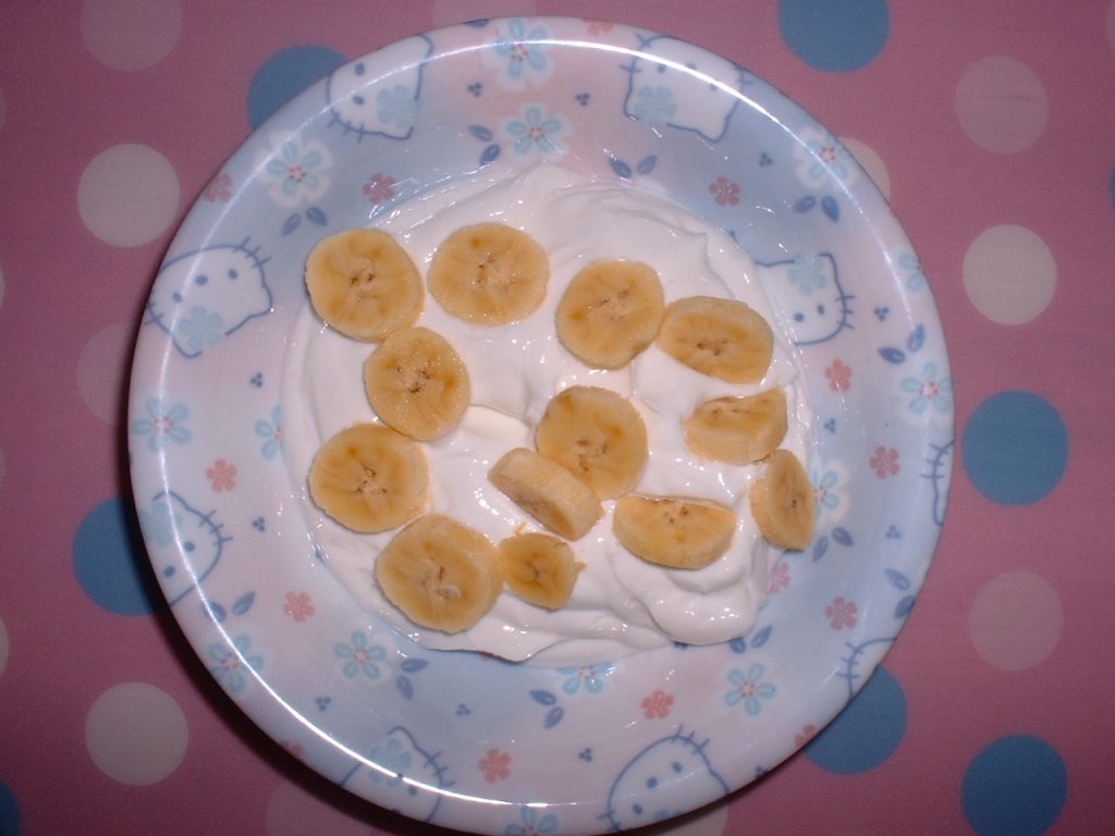 A simple yet inviting breakfast or snack of banana slices and yogurt. The yogurt sits in a clear glass bowl, its smooth, creamy surface a pristine white. The banana slices, in various shades of yellow and flecked with brown at the edges, are nestled into the cool yogurt, their arrangement resembling a half-submerged crescent moon.A single banana slice rests atop the yogurt, its cut end revealing the fruit's soft, creamy flesh. This topmost slice seems to beckon the viewer, inviting them to take a bite and experience the contrasting textures of the chilled yogurt and the yielding banana. The bowl sits on a pastel pink table, the color adding a touch of whimsy to the otherwise minimalist scene. The tabletop itself is smooth and unblemished, reflecting the soft morning light that bathes the composition. To the left of the bowl, a white paper napkin lies crumpled, its folds hinting at the hand that recently reached for a spoonful of the yogurt-banana mix. This small detail adds a sense of intimacy and immediacy to the scene, as if we have just interrupted a moment of quiet enjoyment. Overall, the image evokes a sense of freshness, simplicity, and quiet pleasure. The uncluttered composition and soft colors create a sense of tranquility, while the combination of yogurt and banana suggests a healthy and satisfying start to the day.