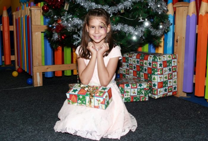 An 11-year-old girl wearing a white dress smiles while holding a gift and sitting on the carpeted floor under the Christmas tree. 