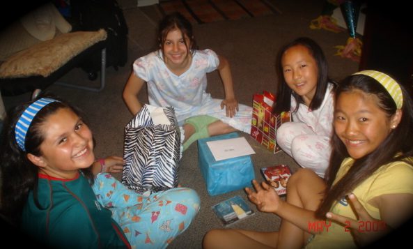 Four girls sit on the floor and smile while opening their gifts.
