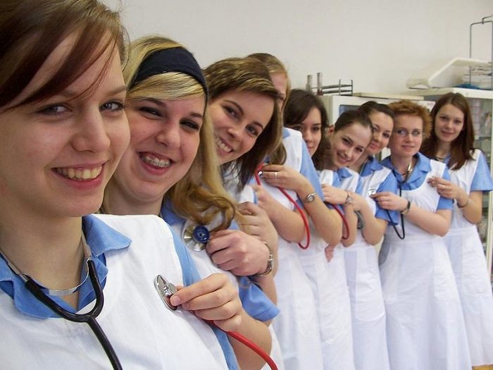 A group of smiling nursing students in uniforms and lab coats, wearing stethoscopes around their necks, posing for a photo in a clinical setting.