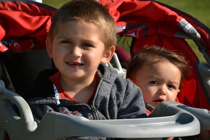 There are two kids in a double carrier. They look so cute. The first child is smiling while the other child looks surprised. 