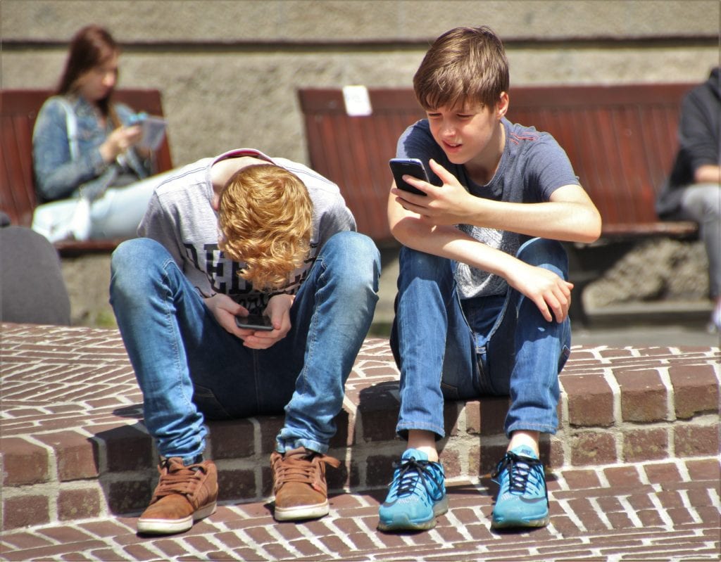 Two young lads sitting on the ladder outdoors while holding their cellphones on a bright day. 