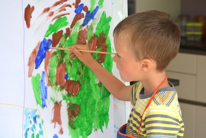 A boy wearing striped shirt and blue apron does his best painting seriously while standing in a brightly lit room. 