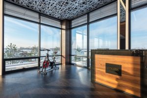 An image of a bright and airy modern home gym with floor-to-ceiling windows offering an expansive city view. A sleek stationary exercise bike, potentially one of the best exercise bikes under $200, stands prominently in the space near a wooden reception desk with a modern design. The room is adorned with an intricate geometric ceiling pattern that adds an artistic touch to the fitness area.