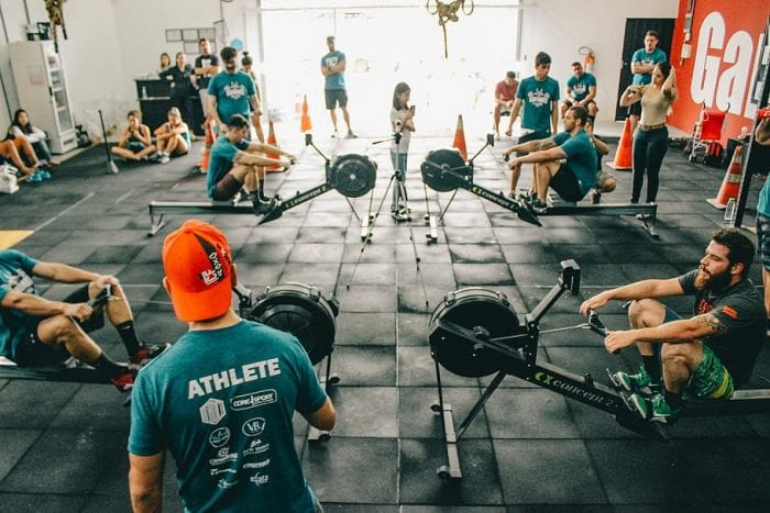 A gym with many people wearing blue-green shirt trying to exercise using the rowing equipment.