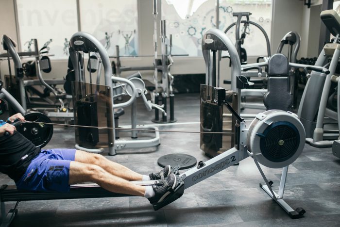 A man with back shirt, blue shorts, and black shoes is inside a gym while doing his best to row the best rowing machine