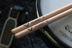 Two 'LOS ANGELES - 5A' drum sticks, ideal as the best drumsticks for beginners, placed on a snare drum, with a foot pedal in the background, indicative of a drumming setup.