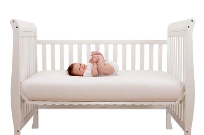 A baby playing with her feet while on a white, comfy crib with mattress.