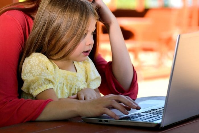 A mother wearing red sweater setting with her child wearing yellow dress facing in front of a gray PC.