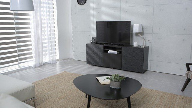 A big home entertainment home area. The design of the home is simple. The white is the most dominant color of the room. There is a black coffee table at the center. There is a big black TV beside the wall.