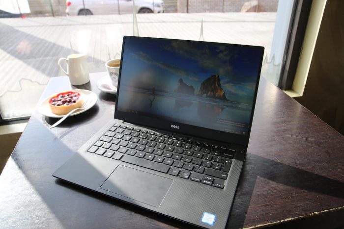 This is Dell best touchscreen laptop. The laptop is on the top of the table. The laptop design is carbon. There is a cheesecake and coffee and creamer on the side. It looks like this picture was taken in the coffee shop. 