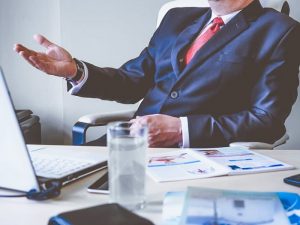 A man is wearing the a suit. It looks like he is explaining something while using his best business laptop.