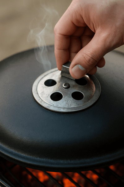 Hand adjusting charcoal grill vent, smoke rising, with glowing coals visible through grate.