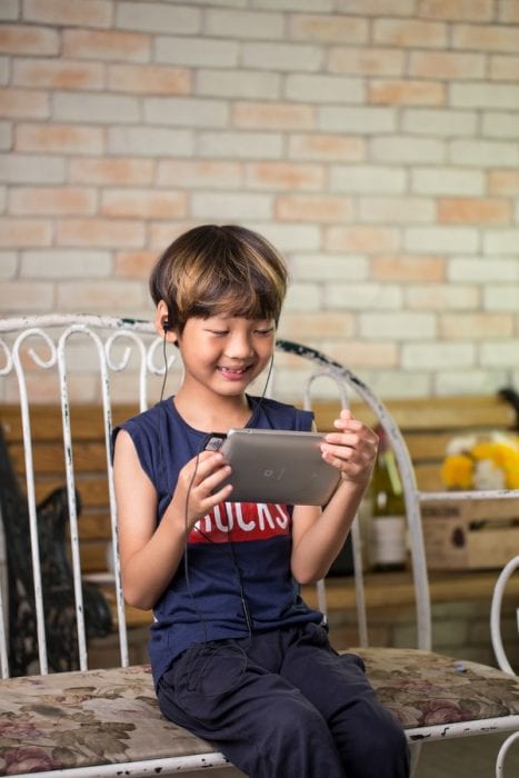 A cute kid sitting on a bench smiling and having fun playing using his tablet for entertainment.