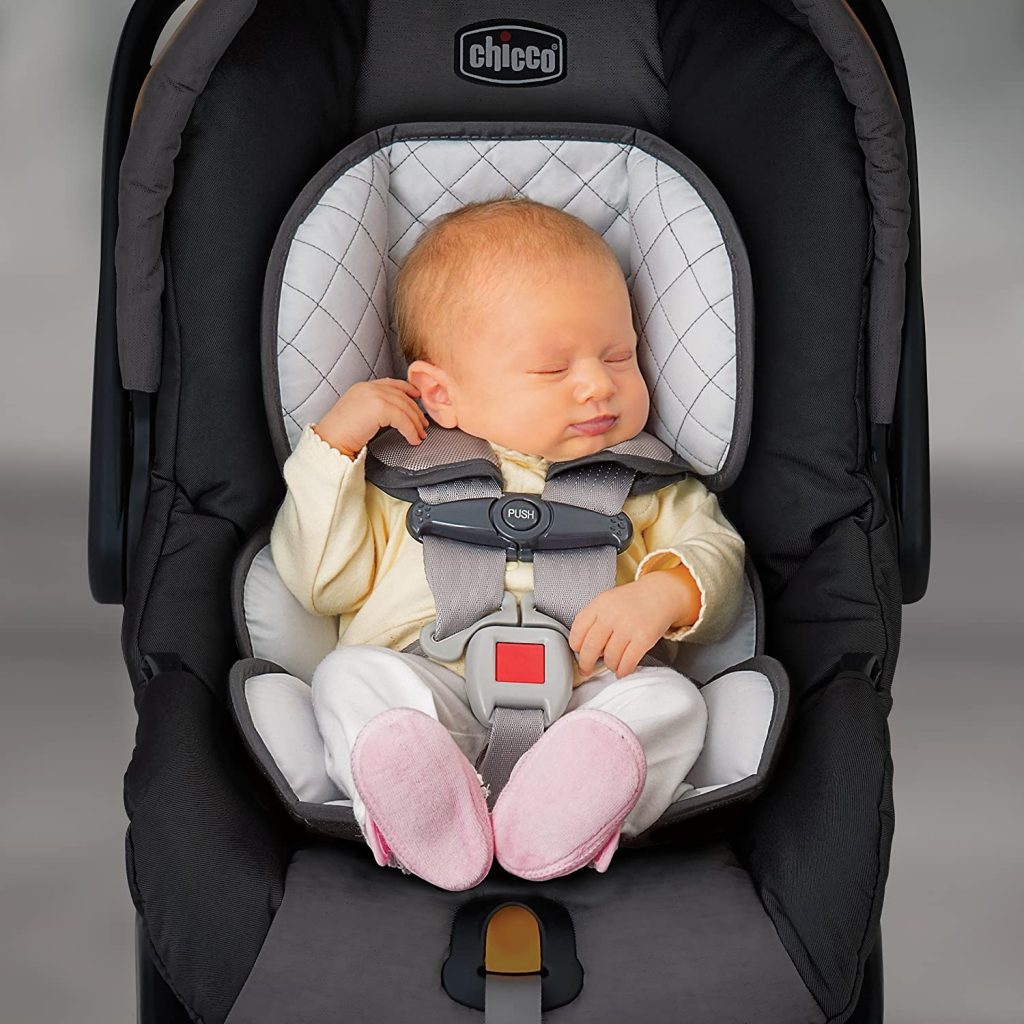 Best car seat for an infant: An infant, peacefully sleeping, securely fastened in a Chicco-brand car seat with a padded five-point harness, a soft insert for extra head support, dressed in a best yellow top, white pants, and pink booties. Perfect infant car seat.