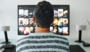 A person wearing a striped shirt sits on the couch and experiences finest sound quality while watching a movie from a television in a bright room.