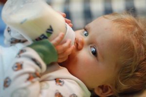 A little kid wearing long sleeves drinks his formula milk while independently holding his bottle of formula. 