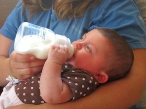 A mother wearing a blue shirt is holding her baby while feeding him with a formula milk from the bottle. 