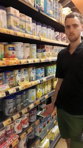 A man wearing a black shirt is in the grocery store, looking for a specific baby formula brand among the many options of baby formulas arranged in the shelves. 
