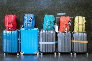 The image shows a row of suitcases and summertime backpacks sitting on top of each other. The summertime suitcases are all different colors and sizes, and the summertime backpacks are all different shapes and sizes. Some of the suitcases have wheels, while others do not. Some of the summertime backpacks have straps, while others do not. There is also a blue duffel summertime bag and a black purse in the image. The summertime suitcases and summertime backpacks are all sitting on a gray tile floor. The background is white, which makes the summertime luggage stand out. There is a plant in the background, and a window with a curtain.