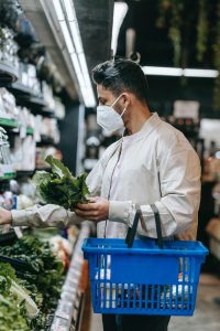 A man shopping at the supermarket for vegetables with vitamin K. He is holding a basket on his left arm, while also holding a leafy vegetable. 