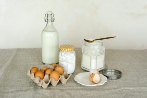 A selection of baking ingredients on a table: a glass bottle of milk, a carton of brown eggs, a jar of sugar with a golden spoon, and a small dish with an egg shell. The items are arranged on a neutral-colored cloth.