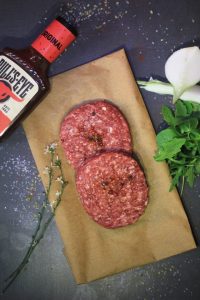 Two raw White Castle burger patties on parchment paper with barbecue sauce, sprigs of thyme, fresh greens, and a cut onion on a dark countertop. These White Castle meat are ready to be place in an air fryer.