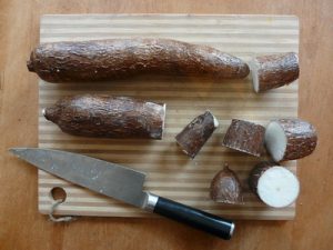 A cassava root resting on a wooden chopping board next to a gleaming, sharp knife, ready to be fry as cassava fries. The cassava is ready for frying.